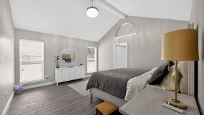 A bedroom with a sloped ceiling, a bed with a gray blanket, a bedside table with a lamp, a dresser with a mirror, and a window with blinds. - Relaxing Retreat Near Lake Lavon (Wylie)