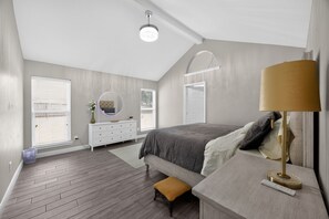Ein Schlafzimmer mit schrägen Wänden, einem Bett mit grauer Decke, einem Nachttisch mit Lampe, einer Kommode mit Spiegel und einem Fenster mit Jalousien.