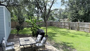 Ein Garten mit Terrassentisch und -stühlen, einem überdachten Grill, einem Holzzaun und einem Baum.