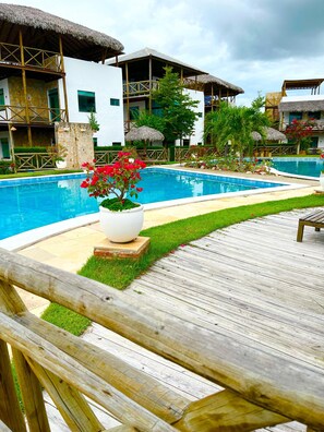 A pool area with a wooden deck, a white building with a balcony, and a thatched-roof structure. - Chaleville Barrinha (Cajueiro da Praia)