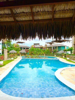 A swimming pool with a tiled edge, surrounded by a garden with potted plants and a thatched-roof structure overhead. - Chaleville Barrinha (Cajueiro da Praia)
