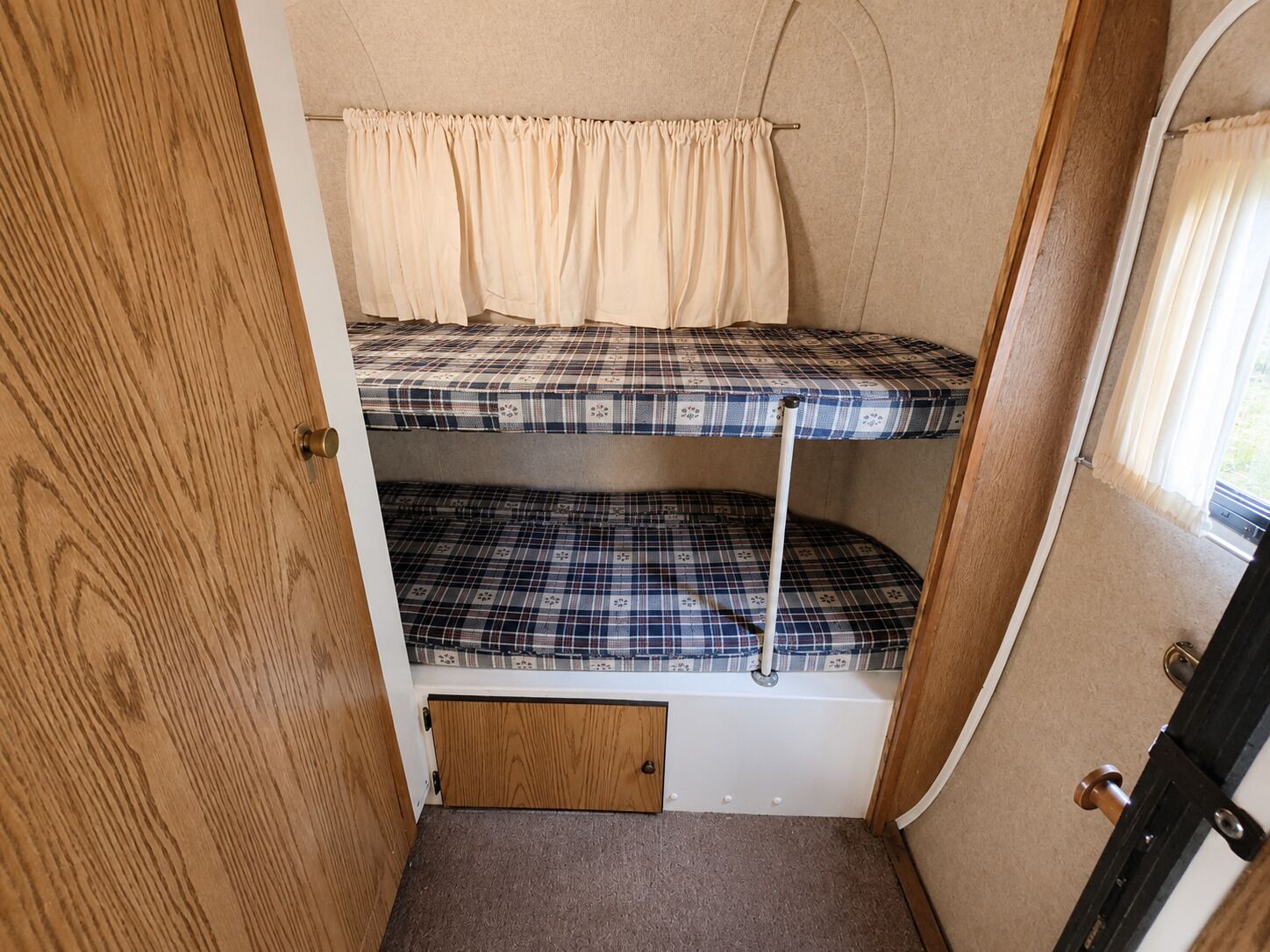 A compact bunk bed with a checkered mattress pattern, a small wooden door, and a window with curtains.