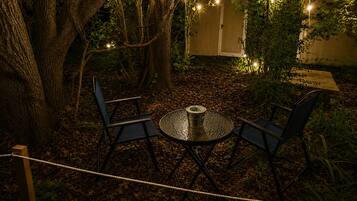 An outdoor seating area with a round table and two chairs, surrounded by trees and string lights.