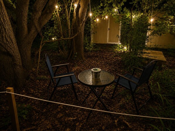 An outdoor seating area with a round table and two chairs, surrounded by trees and string lights.