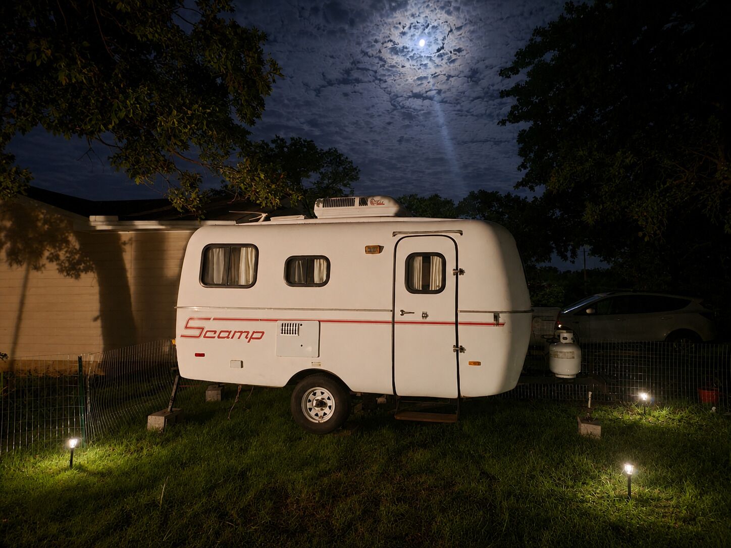 A Scamp RV parked in a grassy area at night with a full moon in the background.