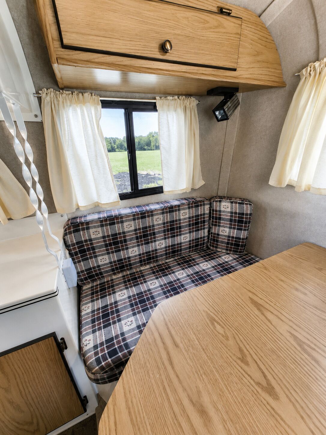 Interior compacto de autocaravana con un asiento de banco a cuadros, una mesita y una ventana con cortinas.