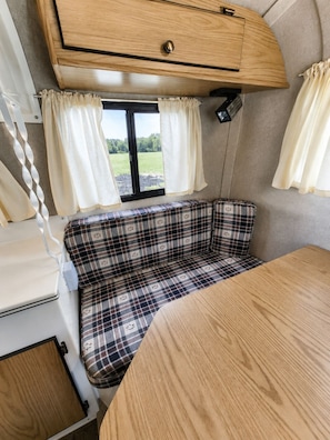 A compact RV interior with a plaid bench seat, a small table, and a window with curtains.