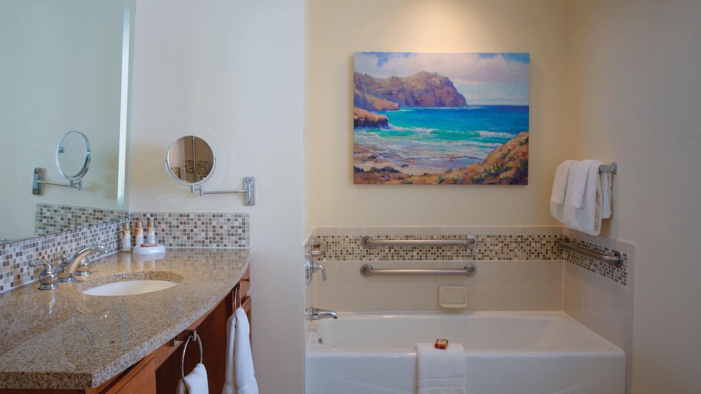 A bathroom with a double sink, a bathtub, a handrail, and a painting of a coastal scene.