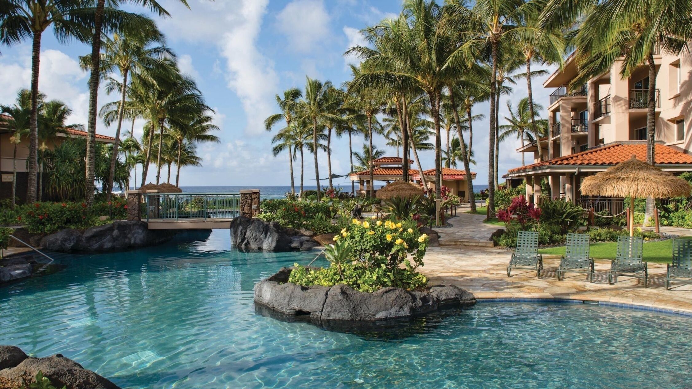 A resort pool with clear water, surrounded by palm trees and a building with a red-tiled roof.
