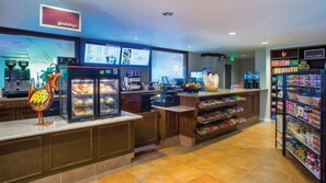 Interior de una cafetería con un letrero de Starbucks, un tablero de menú, una caja registradora, un refrigerador con pasteles y estantes llenos de bocadillos.