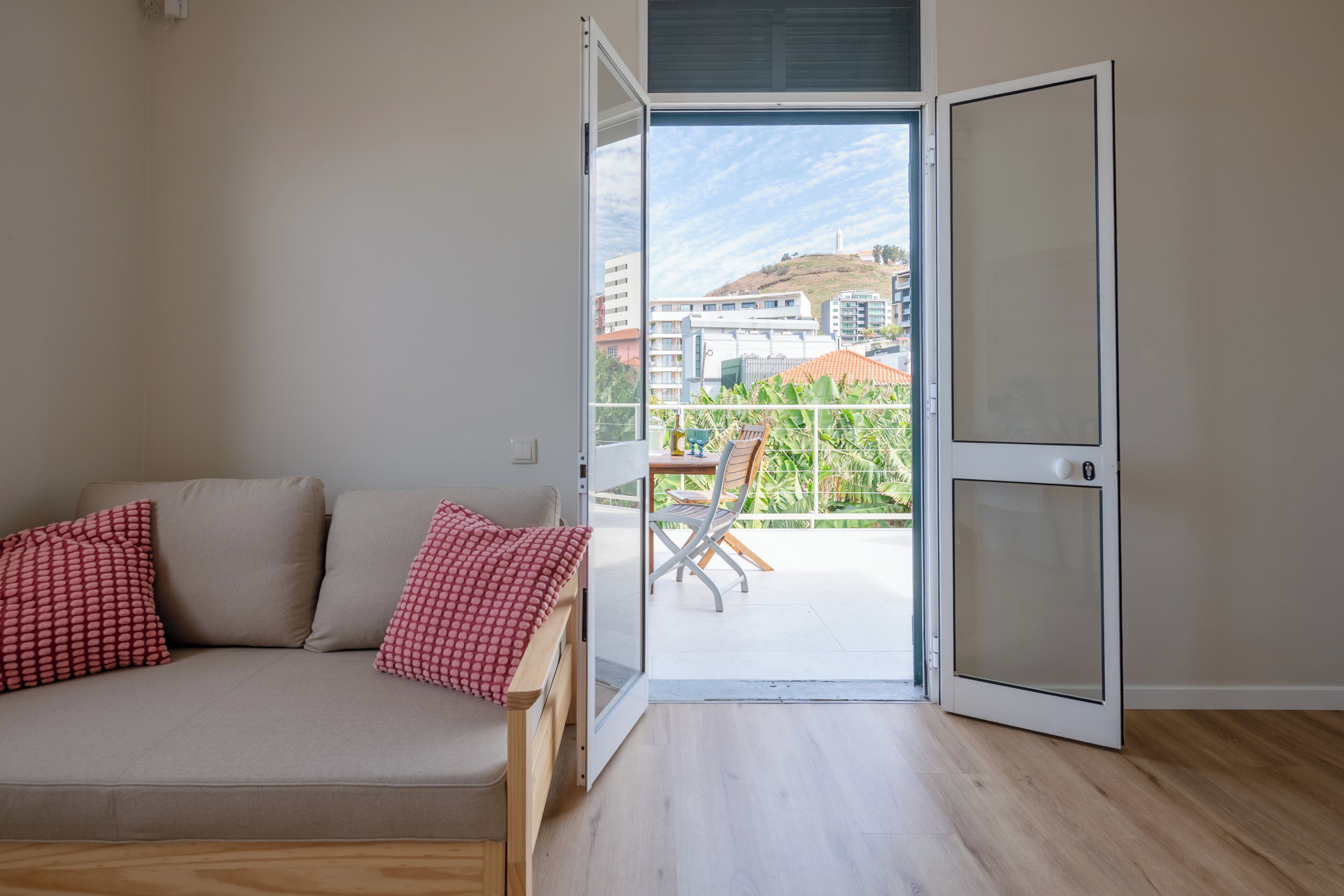 Ein Zimmer mit einer Couch, einer Schiebetür zu einem Balkon mit Blick auf Gebäude und eine Hügelkulisse.