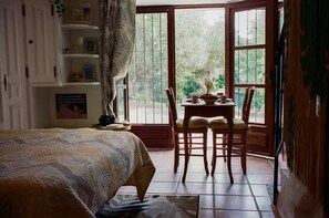 Een slaapkamer met een bed, een tafel met stoelen, een raam met uitzicht op groen en een plank met decoratieve objecten.
