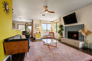 A living room with a fireplace, a ceiling fan, and a television mounted on the wall. - Chromocamino · Pool, Hot Tub, Mini Putt, Game Room (Yucca Valley)
