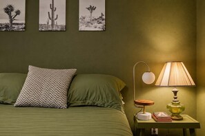 A bedroom with a bed, pillows, a nightstand, a lamp, and wall art. - Chromocamino · Pool, Hot Tub, Mini Putt, Game Room (Yucca Valley)