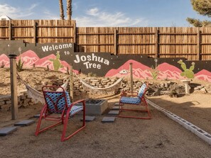 Een buitenruimte in woestijnstijl met twee rode stoelen en een hangmat, een bord met de tekst 'Welcome to Joshua Tree', en een houten omheining op de achtergrond.