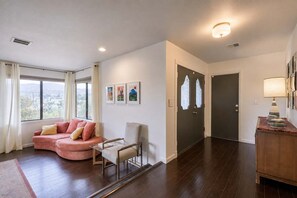 A modern living room with a pink sofa, a glass coffee table, and a chair. There are framed pictures on the wall, a door with frosted glass panels, and a lamp on a wooden side table. - Chromocamino · Pool, Hot Tub, Mini Putt, Game Room (Yucca Valley)