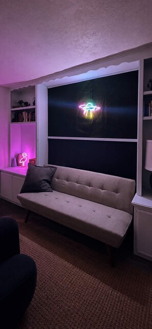 A room with a sofa, a bookshelf, and a neon UFO decoration. - Chromocamino · Pool, Hot Tub, Mini Putt, Game Room (Yucca Valley)