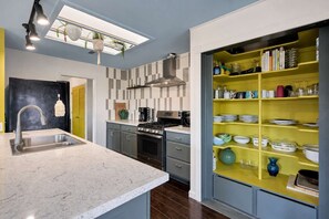 A modern kitchen with a central island, a built-in stove, and a yellow shelving unit filled with dishes. - Chromocamino · Pool, Hot Tub, Mini Putt, Game Room (Yucca Valley)