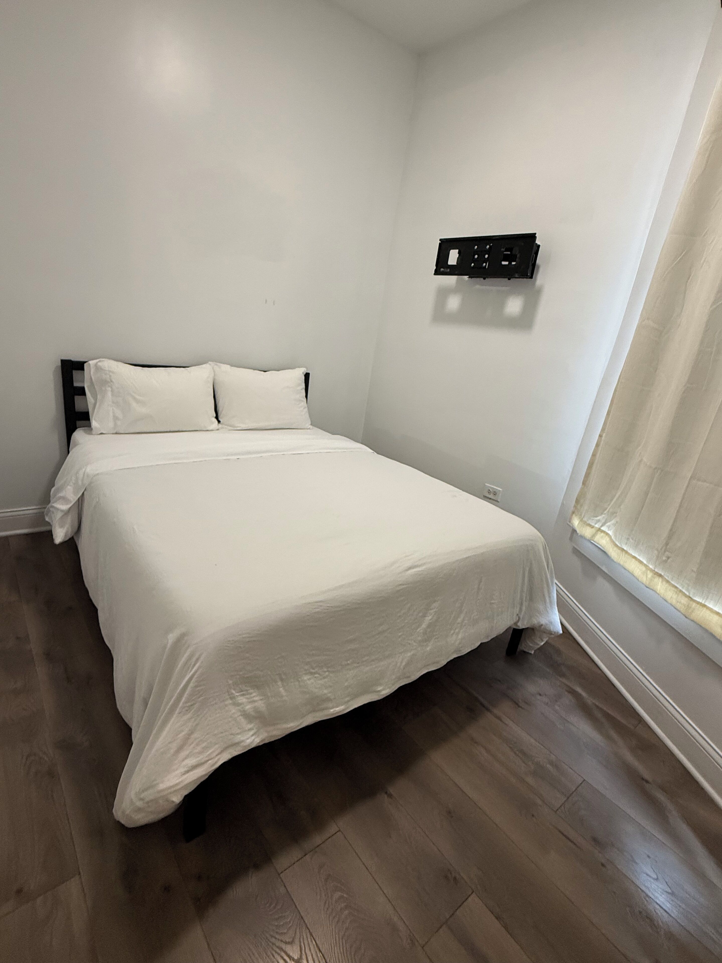 Une chambre à coucher avec un lit, des murs blancs, une fenêtre avec des rideaux et une prise électrique fixée au mur.