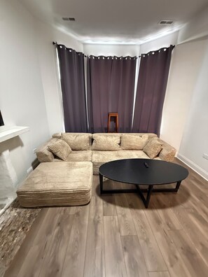 Un salón con forma de esquina, un sofá modular, una mesita redonda y un taburete de madera.