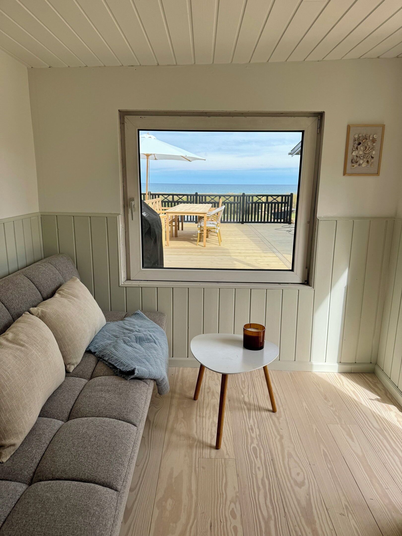 A room with a view of a beach, a wooden bench, a table with a candle, and a grey sofa.