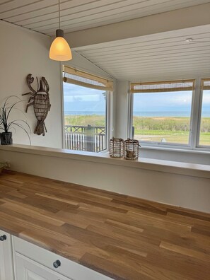 A kitchen with a wooden countertop, a hanging light fixture, and a view of the beach through large windows.