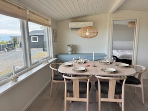 A dining area with a round table set for six, wooden chairs, a hanging light fixture, and a window with a view of the outside.