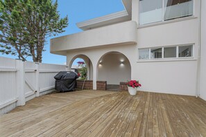 Eine Holzterrasse mit Grill, weißem Zaun und einem Kübel mit rosa Blüten.
