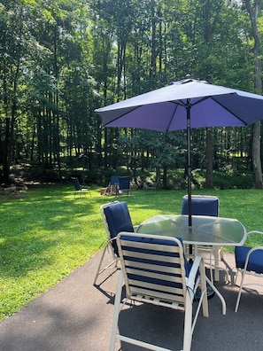 An outdoor patio with a table and chairs, a purple umbrella, and a grassy area surrounded by trees.