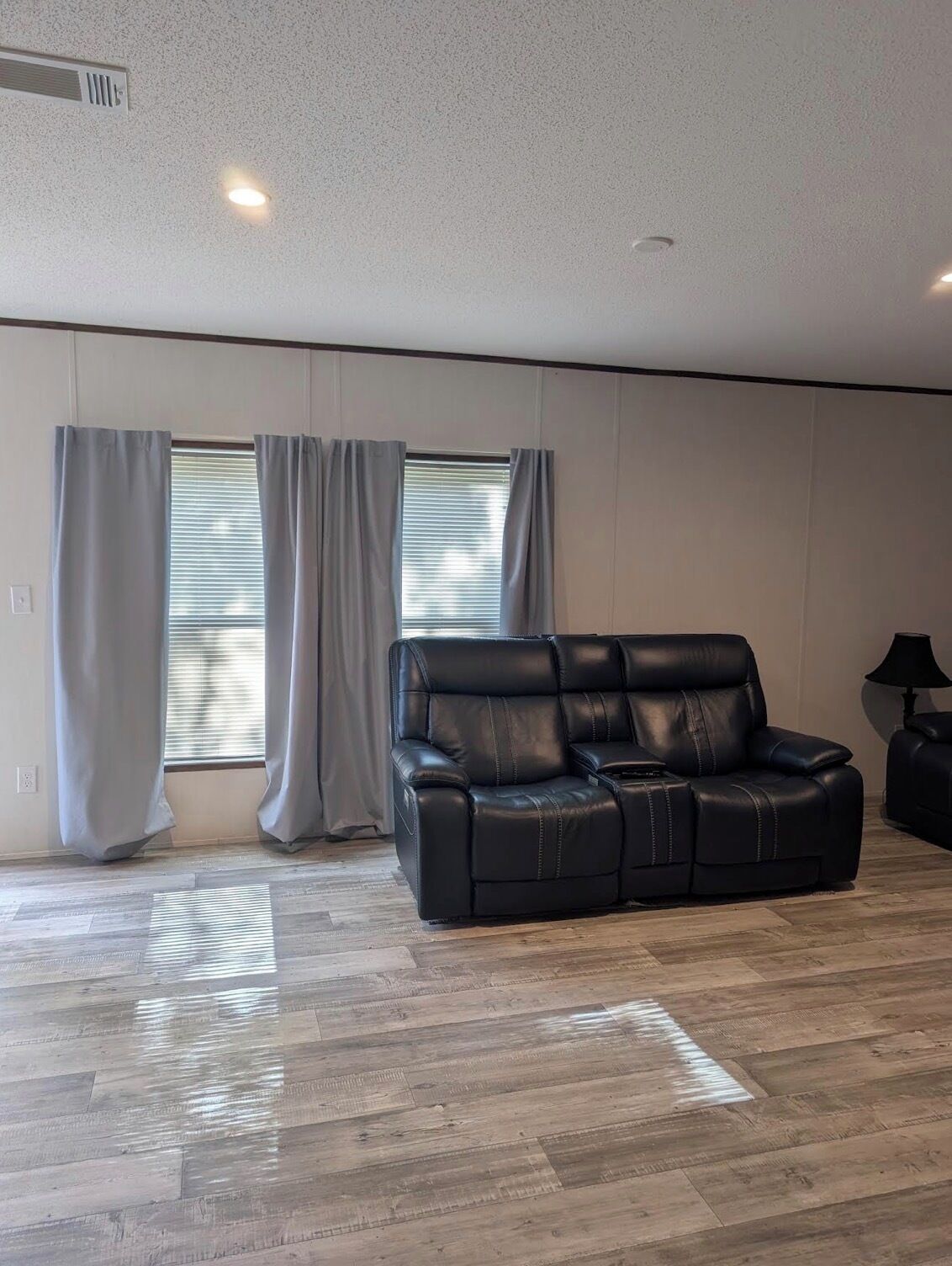 A spacious living room with a black leather sofa, wooden flooring, and a window with blinds.
