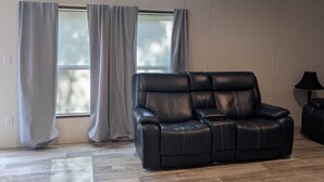 A spacious living room with a black leather sofa, wooden flooring, and a window with blinds.
