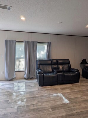 A spacious living room with a black leather sofa, wooden flooring, and a window with blinds.
