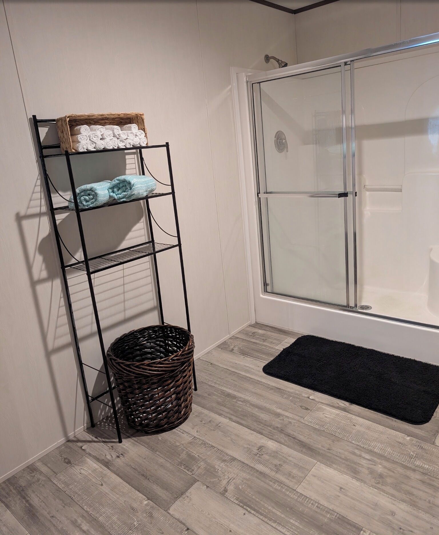 A bathroom with a shower, a towel rack with folded towels, a wicker basket, and a black doormat.