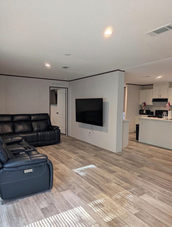 A modern living room with a black leather sofa, a flat-screen TV mounted on the wall, and a kitchen area in the background.