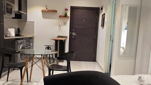 Ein modernes Wohnzimmer mit einer dunklen Couch, einem Essbereich mit Tisch und Stühlen, einer Küche mit Geräten und einer dunklen Tür.