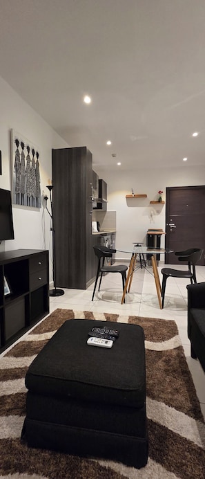 A modern living room with a sofa, a coffee table, and a TV. There is a kitchen area with a fridge, microwave, and a small dining table. - ebs properties 2 two bedroom duplex (Accra)
