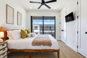 A bedroom with a bed, bedside table, lamp, ceiling fan, and a TV mounted on the wall. - Unwind in Style: Luxe Condo Getaway! (New Braunfels)