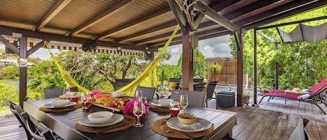 Un área de comedor al aire libre con una mesa de madera preparada para una comida, rodeada de sillas y un espacio de estar cubierto con una hamaca.