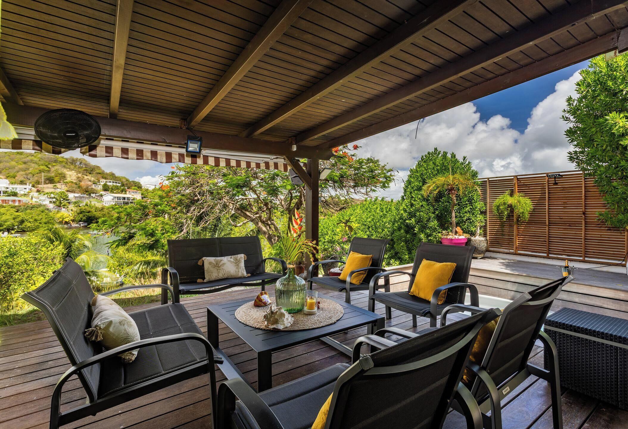 Un patio al aire libre con cubierta de madera, sillas de metal negro, una mesa con posavasos tejido y vistas a una ladera con vegetación y edificios.