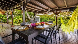 An outdoor dining area with a wooden table set for two, surrounded by chairs, under a wooden pergola with a hammock. - Beachfront Villa - Private Paradise (Le Robert)