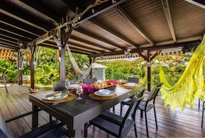 Ein Essbereich im Freien mit einem Holztisch, gedeckt für zwei Personen, umgeben von Stühlen, unter einer Holzpergola mit einer Hängematte.