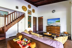 A living room with a wooden staircase, a sofa with patterned cushions, a television on a wooden cabinet, and a beach scene painting on the wall. - Beachfront Villa - Private Paradise (Le Robert)