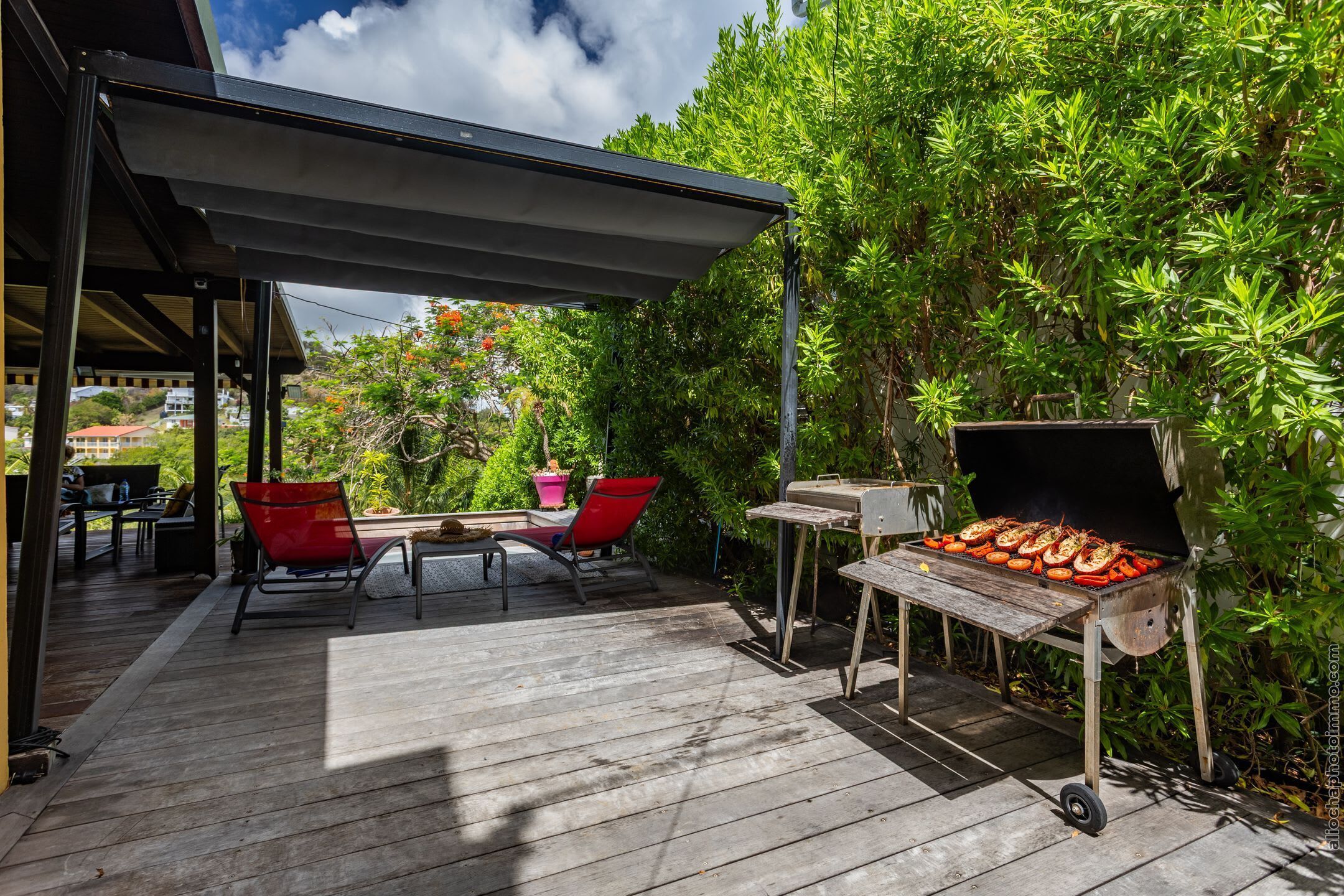 A wooden deck with a barbecue grill, red chairs, and a covered patio area.