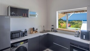 Ett modernt kök med inbyggd mikrovågsugn, kaffebryggare och mixer. Köket har ett stort fönster med utsikt över havet och frodig grönska.