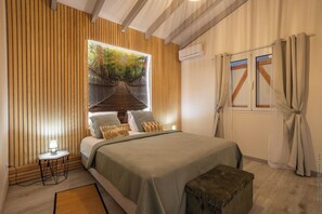 Ein Schlafzimmer mit einem Bett, einem Nachttisch, einer Lampe, einem Fenster mit Vorhängen und Blick auf einen Wald durch ein Insektengitterfenster.