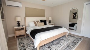 Un dormitorio con cama, mesitas de noche, ventilador de techo, aire acondicionado, alfombra estampada y un espejo.
