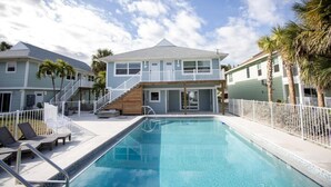 Una piscina con valla blanca, sillas de descanso y un edificio de dos pisos con balcón.