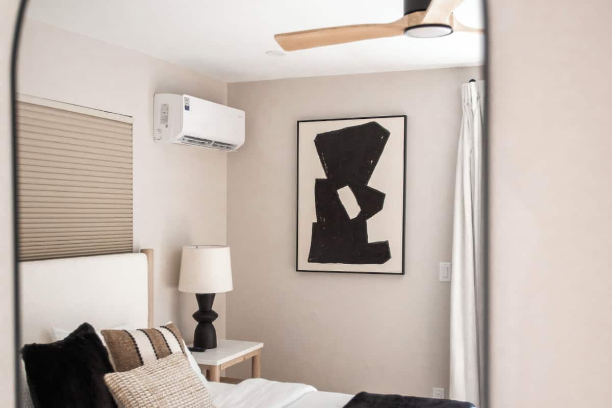 Un dormitorio con cama, mesita de noche, lámpara, unidad de aire acondicionado y una obra de arte enmarcada en la pared.