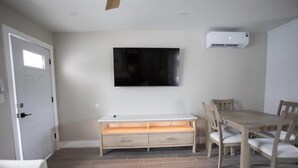 Una habitación con una televisión blanca montada en la pared, una mesa de comedor de madera con sillas, un banco blanco con cajones y una alfombra estampada en el suelo.