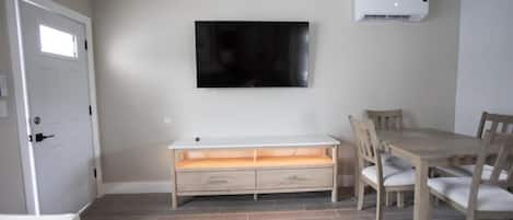 Una habitación con una televisión blanca montada en la pared, una mesa de comedor de madera con sillas, un banco blanco con cajones y una alfombra estampada en el suelo.